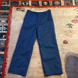 Blå byxor från Carhartt - Snygga blå byxor från Carhartt med en klassisk rak passform. Byxorna har en midwaist och är perfekta för en stilren look. De är tillverkade i ett slitstarkt material och har en knappstängning framtill.