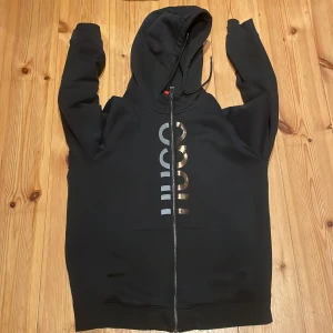 Hugo boss tröja  - Snygg svart hoodie från Hugo med dragkedja och tryck framtill. Perfekt för en casual look med sin bekväma passform och stilrena design. Huvan har justerbara snören för extra komfort. Koftan är strl medium i väldigt bra skick 