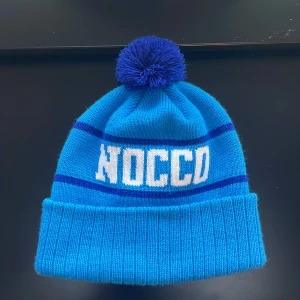 Ljusblå mössa med tofs från NOCCO - Snygg blå mössa från NOCCO med en stor tofs på toppen. Mössan har mörkblå ränder och texten 'NOCCO' i vitt framtill. Perfekt för kalla dagar!