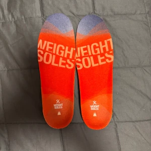 Inläggssulor från Weight Soles - Säljer ett par färgglada inläggssulor från Weight Soles. Ovansidan har en gradient i rött och lila med texten 'Weight Soles'. Undersidan är grå med ett mönster för extra grepp. Perfekta för att ge extra komfort och stöd i dina skor.