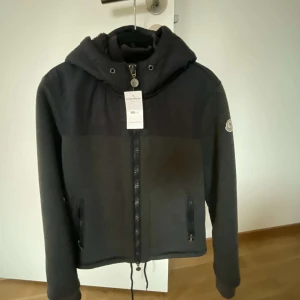 Jacka från Moncler - Snygg jacka från Moncler med huva och dragkedja. Jackan har ett stilrent märke på ärmen och är perfekt för kyligare dagar. Den har en mjuk insida och praktiska fickor med dragkedja. Jackan är köpt från store enda. Kvitto finns