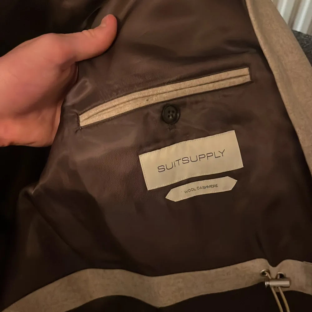Elegant beige kappa från Suitsupply, tillverkad i en lyxig blandning av ull och kashmir. Jackan har en stilren design med knappar och två stora fickor framtill. Perfekt för en sofistikerad look. Ny pris 6,899kr. Takit.