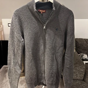 Grå kofta från MCS - Säljer en stilren grå kofta från MCS med dragkedja och ribbad krage. Perfekt för kyligare dagar och enkel att matcha med olika outfits.