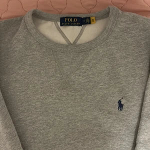 Helt ny grå sweatshirt från Polo Ralph Lauren - Säljer en klassisk grå sweatshirt från Polo Ralph Lauren med rund hals och det ikoniska logotypbroderiet på bröstet. Tröjan har långa ärmar och ribbade muddar vid ärmslut och nederkant. Perfekt för en avslappnad stil.