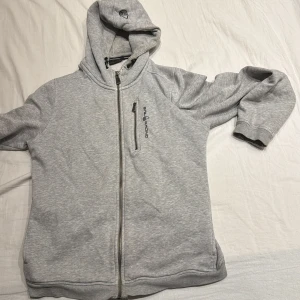 Grå hoodie från Sail Racing - Säljer en grå hoodie från Sail Racing med dragkedja och en bröstficka. Perfekt för kyliga dagar med sin mjuka och bekväma passform. Huvan har justerbara snören för extra komfort.