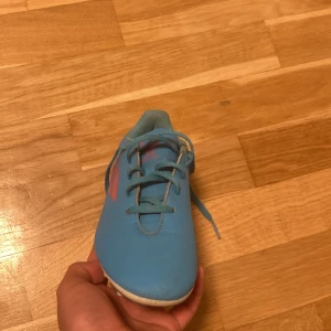 Blå fotbolls dobbar från Adidas - Säljer ett par blå fotbollsskor från Adidas med rosa detaljer och snörning. Skorna har dobbar för bra grepp på planen och är tillverkade i syntetmaterial.