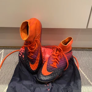 Nike Hypervenom Elite FG - Snygga Nike fotbollsskor i en kombination av orange och svart. Skorna har en hög ankelkrage och snörning för bra passform. Perfekta för fotbollsplanen med sin slitstarka sula och moderna design. Observera det lilla hålet längst fram vid höger stortå på sista bilden.