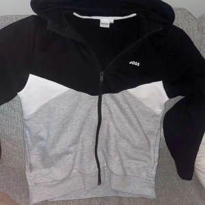 Svart och grå hoodie från Boss - Snygg hoodie från Boss i svart och grått med dragkedja framtill. Den har en justerbar huva och vita detaljer på sidorna. Perfekt för en avslappnad stil.Den har bara använts 2-3 gånger. Ny pris 1499
