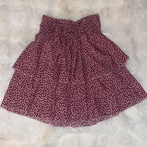 Blommig kortkjol - Söt blommig kortkjol i rosa/lila och vit med elastisk midja. Tillverkad i Italien av polyester och elastan, vilket ger en bekväm passform. Perfekt för varma dagar. Säljer pga att jag tycker själv att den är för lång, vek den alltid när jag använde den. Använd ca 3 gnr.