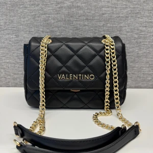 Svart quiltad axelväska från Valentino - Snygg svart quiltad axelväska från Valentino med guldfärgade kedjedetaljer. Väskan har en elegant design med ett lock och en säker stängning. Perfekt för att ge en lyxig touch till din outfit. Knappt använd!