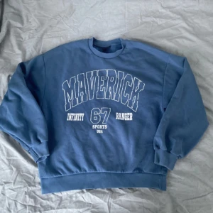 Blå sweatshirt från Gina Tricot - Säljer en blå sweatshirt från Gina Tricot med texten 'Maverick Infinity 67 Ranger Sports USA' tryckt på framsidan. Tröjan har långa ärmar och ribbade muddar vid ärmslut och nederkant. Perfekt för en avslappnad stil.