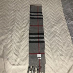 Burberry halsduk - Snygg och lyxig grå halsduk från Burberry i 100% kashmir. Använd 2 gånger. Den har ett klassiskt rutmönster i grått och rött, perfekt för att hålla dig varm och stilren under kyliga dagar.