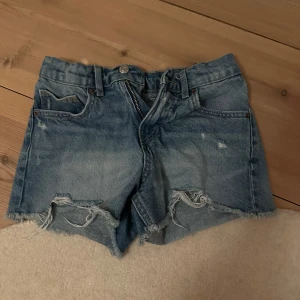 Blå jeansshorts med slitningar - Snygga blå jeansshorts med slitna detaljer och råa kanter. Perfekta för en avslappnad stil. De har klassisk femficksdesign och knäppning med dragkedja.