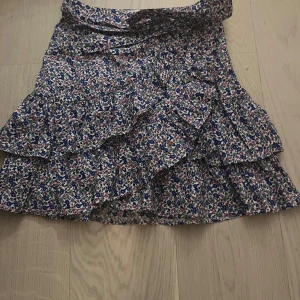 Blommig kjol med volanger - Charmig blommig kjol med volanger i blått, rosa och vitt. Kjolen har en omlott-design och knytband i midjan för en justerbar passform. Perfekt för en somrig look!