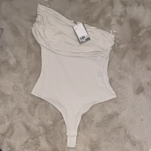 Vit oneshoulder bodysuit - Snygg vit oneshoulder bodysuit med draperad detalj över axeln. Perfekt för en stilren outfit. Tillverkad i ett mjukt och stretchigt material för en bekväm passform.