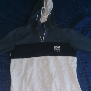 Grå och svart hoodie från Jack & Jones - Säljer en snygg hoodie från Jack & Jones i grått och svart med dragkedja och justerbar huva. Perfekt för en avslappnad stil. Märket 'CORE' syns på bröstet.