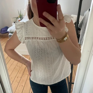 Vit blus med spetsdetaljer - Säljer en söt vit blus med spetsdetaljer och volangärmar. Blusen har ett fint hålmönster och en rak passform. Perfekt för vår och sommar!