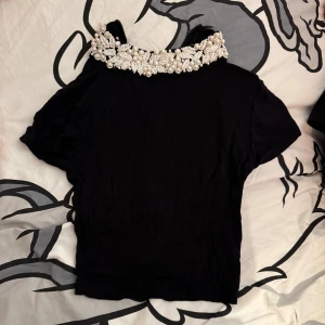 Svart topp med pärldetaljer - Snygg svart topp med korta ärmar och en fantastisk pärlbroderad krage. Toppen har en normal passform och är tillverkad i ett mjukt material.