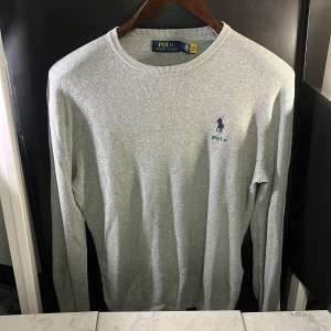 Grå tröja från Polo Ralph Lauren - Säljer en stilren grå tröja från Polo Ralph Lauren med rund halsringning och det klassiska logotypbroderiet i blått på bröstet. Tröjan har långa ärmar och är perfekt för en avslappnad look. Det finns defekter på baksidan av kragen, annars mycket fint skick. Hör av er vid minsta fundering🙌
