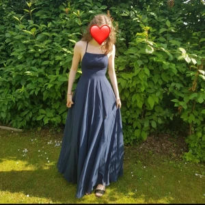 Mörkblå balklänning med öppen rygg - Mörkblå balklänning med en slits framtill. Den har en öppen rygg med snörning. Perfekt längd med klackar för mig som är 172cm. Endast använd en gång, original pris 1486kr. Storlek 32, men passar större också.