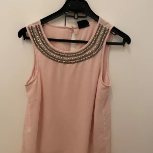 Rosa ärmlös topp med pärldetaljer - Elegant rosa ärmlös topp från Vila med vackra pärldetaljer runt halsringningen. Perfekt för en stilren look.