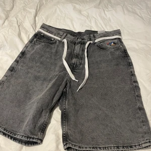 Grå jeansshorts från Cava Company - Säljer ett par grå jeansshorts från Cava Company med vit snörning i midjan. Shortsen har en avslappnad passform och klassiska femficksdetaljer. Perfekta för en casual look.