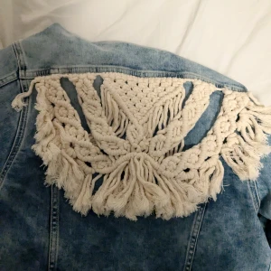 Jeansjacka med virkad detalj från Never Denim - Snygg jeansjacka från Never Denim med unik virkad detalj på ryggen. Jackan har en klassisk blå tvätt och knappar framtill. Perfekt för att ge din outfit en bohemisk touch.