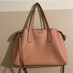 Rosa axelväska från Guess - Snygg rosa axelväska från Guess med stilren design och praktisk dragkedja. Väskan har två handtag och en justerbar axelrem för bekväm användning. Obs den är äkta!! Jag säljer väskan för att jag aldrig använt så den är i mycket bra skick!💓 
