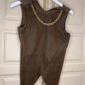 Brun ärmlös klänning med guldkedja - Snygg brun ärmlös klänning med en iögonfallande guldkedja vid halsringningen. Klänningen har en asymmetrisk design och är tillverkad i ett mjukt material som ger en bekväm passform. Perfekt för att ge en elegant touch till din garderob.