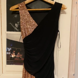 Svart och guldglittrig klänning - Snygg klänning med en unik design i svart och guldglitter. Den har en oneshoulder-stil och en elegant drapering. Perfekt för en kväll ute!