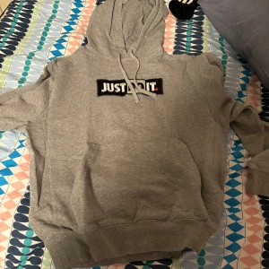 Grå hoodie från Nike - Säljer en grå hoodie från Nike med texten 'Just Do It' på bröstet. Den har en klassisk design med huva och dragsko samt ribbade muddar vid ärmslut och nederkant. Perfekt för en avslappnad stil.