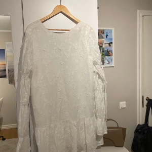 Vit klänning med blommigt mönster - Elegant vit klänning med blommigt mönster och v-ringad design. Klänningen har långa ärmar och en volangdetalj vid fållen. Perfekt för vår och sommar.