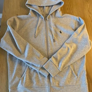 Grå hoodie från Polo Ralph Lauren - Säljer en stilren grå hoodie från Polo Ralph Lauren med dragkedja och broderad logga på bröstet. Perfekt för en avslappnad look. Den har en klassisk passform och är tillverkad i mjukt material.