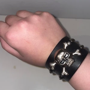 Svart armband med dödskalle och nitar - Coolt svart armband med en stor silverfärgad dödskalle och korslagda ben. Armbandet har flera nitar och ett justerbart spänne.