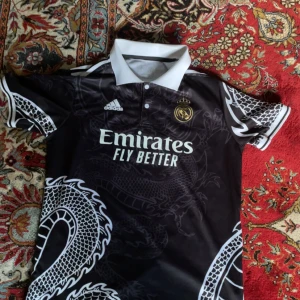 Svart Real Madrid fotbollströja - Snygg svart Real Madrid fotbollströja med drakmönster och vit krage. Tröjan har korta ärmar och klubbens emblem i guld på bröstet. Benzema och nummer 9 pryder baksidan. Perfekt för fans av laget!