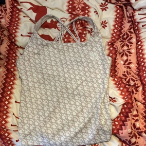 Beige mönstrad topp från Guess - Snygg beige topp från Guess med ett stilrent mönster och korsade axelband. Perfekt för en trendig look. Märkeslogga på framsidan ger en extra touch av stil. (Känns som baddräkts material)