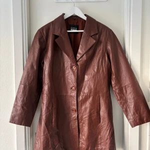 Brun skinnkappa från Steve - Snygg brun skinnkappa från Steve med knappar framtill. Kappan har en klassisk krage och långa ärmar, perfekt för höst och vår. Tillverkad i äkta läder. Diskreta fickor på sidorna. Mindre repa, se bild. Strl saknas men bedöms som M.