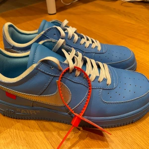 Blå Nike Air Force med vit snörning - Snygga blå Nike Air Force med vit snörning och silverdetaljer. Skorna har en röd tagg och textdetaljer på sidan. Perfekta för en stilren look.