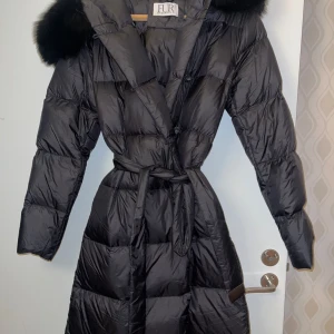 Pälsjacka från Fur Stockholm - Snygg svart dunjacka från Fur Stockholm med pälsdetaljer på huvan. Jackan har ett bälte i midjan och knäppning framtill. Perfekt för kalla dagar med sin långa design och vadderade stil. Endast använd fåtal gånger då det inte är min stil riktigt. Dock väldigt fin jacka och i nyskick. 