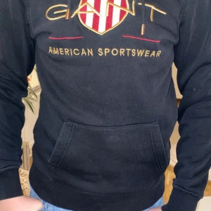 Svart hoodie från GANT - Säljer en snygg svart hoodie från GANT med deras klassiska logga i rött och guld på bröstet. Den har en känguruficka framtill och är perfekt för en casual look. Passar bra till både jeans och joggers!