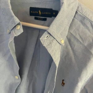 Blå skjorta från Ralph Lauren - Säljer en snygg blå skjorta från Ralph Lauren i storlek M. Skjortan är långärmad och har en klassisk krage med knappar. Den är tillverkad i bomull och har en liten broderad logga på bröstet. Perfekt för både vardag och fest!