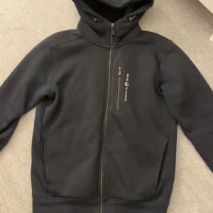 Svart hoodie från Sail Racing - Säljer en svart hoodie från Sail Racing i storlek S. Den har en dragkedja framtill och en praktisk huva. Perfekt för kyliga dagar och har en stilren design med Sail Racing-loggan på bröstet. Materialet är en blandning av bomull och polyester vilket gör den både bekväm och hållbar.