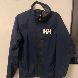 Blå vindjacka från Helly Hansen - Säljer en snygg blå vindjacka från Helly Hansen i storlek S. Jackan har en dragkedja framtill och är långärmad med Helly Hansen-logga på bröstet och ärmen. Perfekt för blåsiga dagar och har en stilren design.
