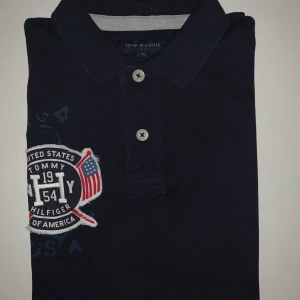 Navy pikétröja från Tommy Hilfiger - Säljer en marinblå pikétröja från Tommy Hilfiger i storlek M. Tröjan har en broderad logga på bröstet och knappar framtill. Perfekt för en stilren och avslappnad look. Passformen är slim fit.