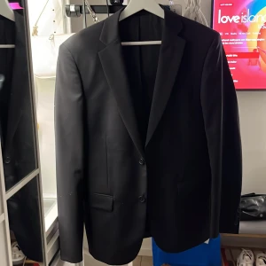 Svart kavaj från Dressmann - Säljer en stilren svart kavaj från Dressmann i storlek S. Kavajen har en klassisk design med två knappar framtill och fickor på sidorna. Insidan har snygga röda detaljer som ger en extra touch. Perfekt för både fest och mer formella tillfällen! Skriv