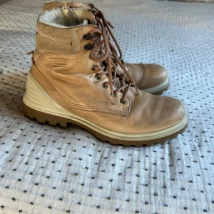 Beige kängor från Ecco - Säljer ett par robusta och snygga beige kängor från Ecco. De är i skinn och har en rejäl sula som passar perfekt för höst och vinter. Kängorna har snörning och dragkedja för enkel på- och avtagning. Insidan är fodrad för extra värme och komfort. Perfekta för kalla dagar!