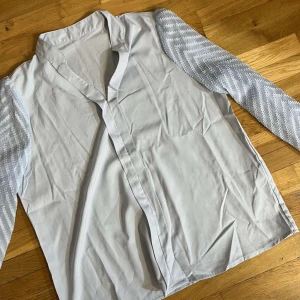 Vit blus med mönstrade ärmar - Säljer en stilren vit blus med unika mönstrade ärmar i ljusblått och vitt. Blusen har en v-ringad design och den har långa ärmar med ett snyggt zigzag-mönster, den är i storlek L men sitter som M, den är helt ny utan defekter