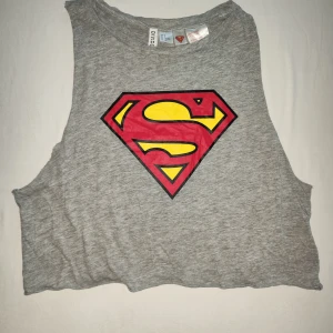 Grå Superman crop top linne från Divided - Säljer ett grått linne från Divided med det ikoniska Superman-logotypen i rött och gult på framsidan. Perfekt för en avslappnad look eller som en cool detalj till din outfit. Linnet är ärmlöst och har en bekväm passform. Perfekt för sommarens varma dagar! 🦸‍♂️