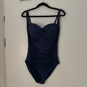 Mörkblå baddräkt - Säljer en elegant mörkblå baddräkt med en stilren design. Den har en vacker rynkad detalj framtill och justerbara axelband för bästa passform. Perfekt för en dag på stranden eller vid poolen! Oklart om storlek men använts av en som bär M annars 