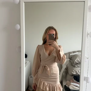 Beige omlottklänning med volangdetaljer - Säljer en superfin beige/ ljusrosa omlottklänning med volangdetaljer. Klänningen har långa ärmar och ett knytband i midjan 🤍 andvänd 1 gång